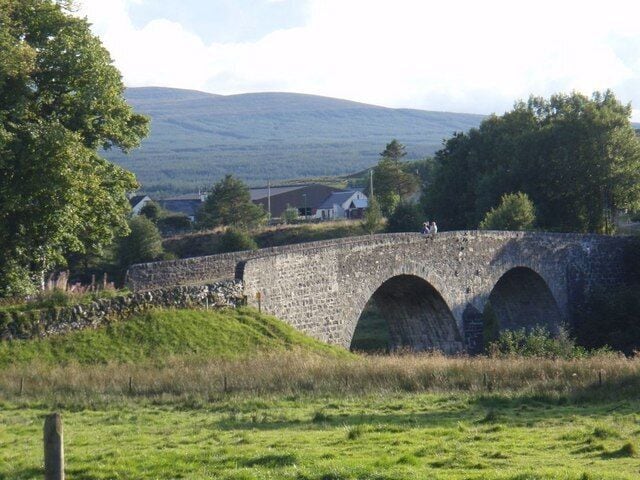 A stone bridge with three arches over a grassy field, surrounded by trees and hills in the background.