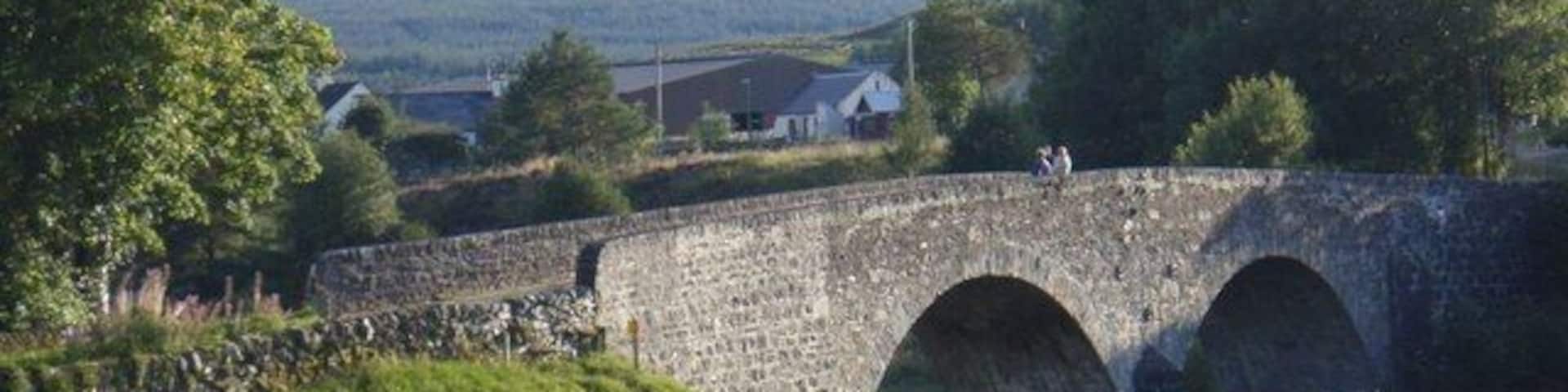 A stone bridge with three arches over a grassy field, surrounded by trees and hills in the background.