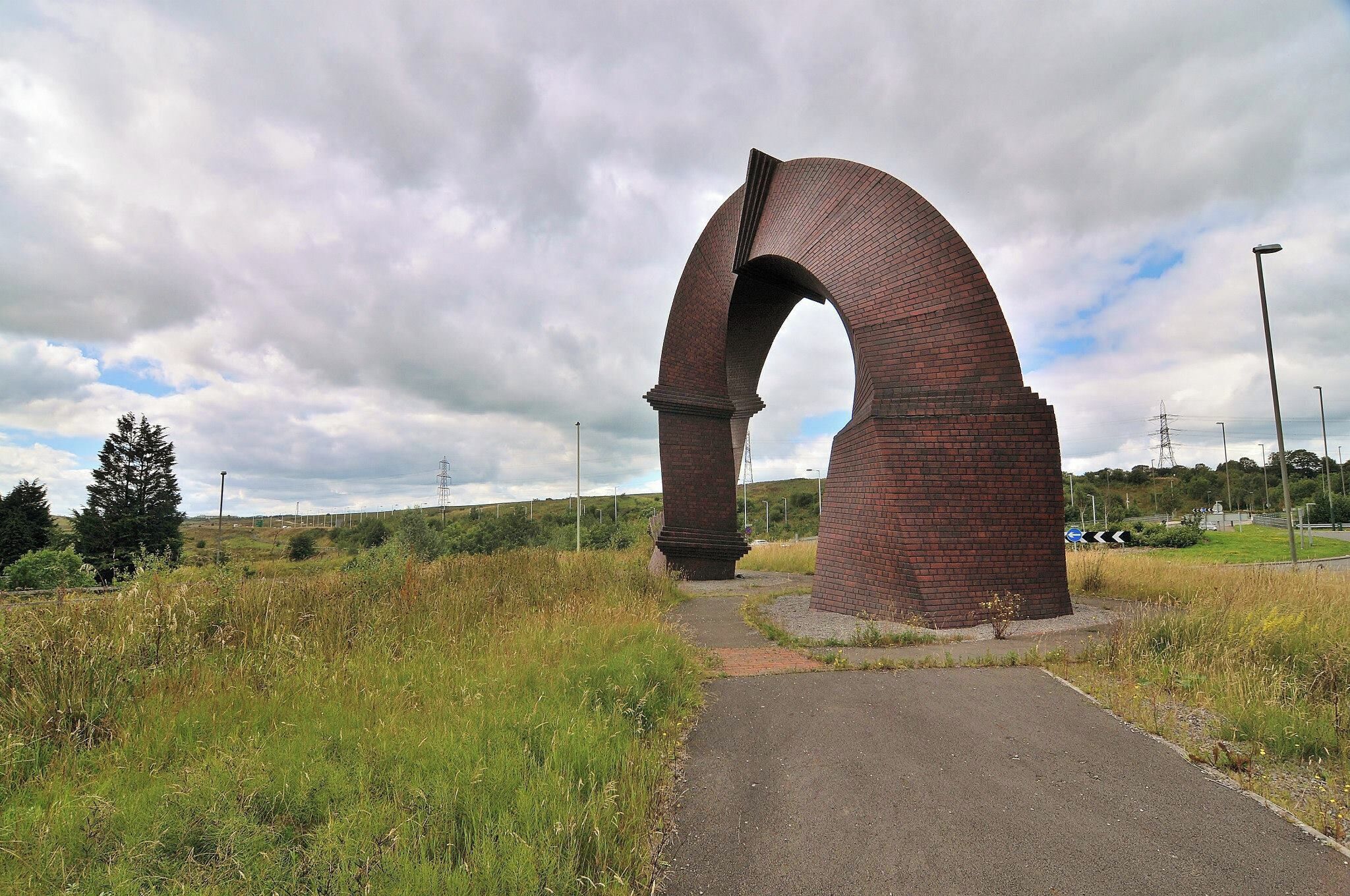 A large brick archway in a grassy field with a paved path leading through it.
