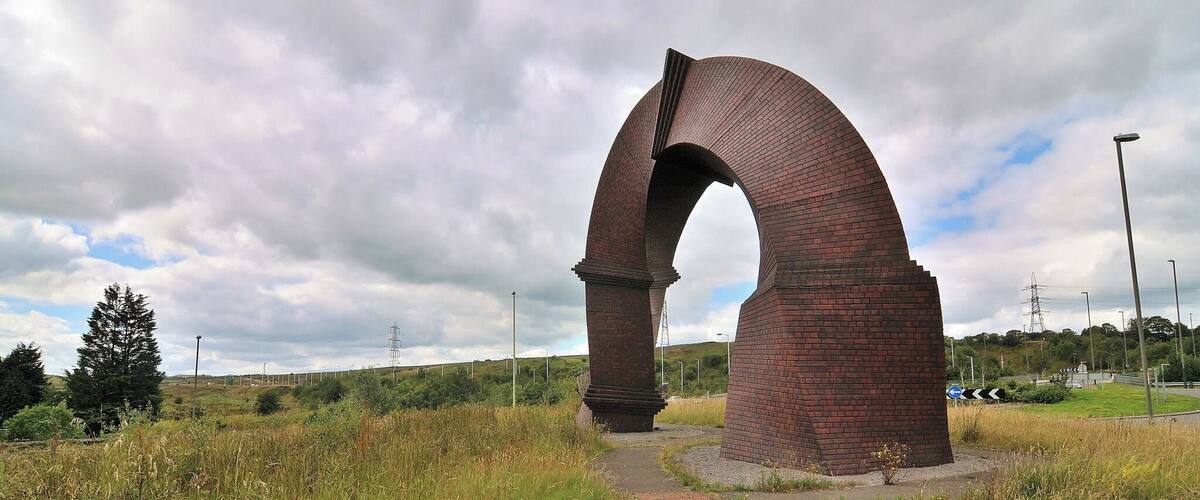 A large brick archway in a grassy field with a paved path leading through it.
