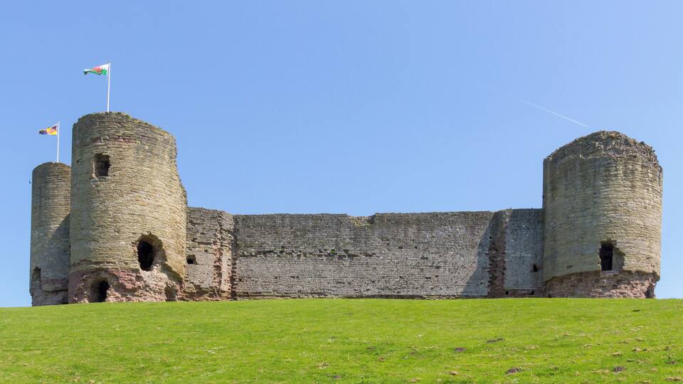 A historic stone castle with two round towers, a grassy foreground, and a clear blue sky.