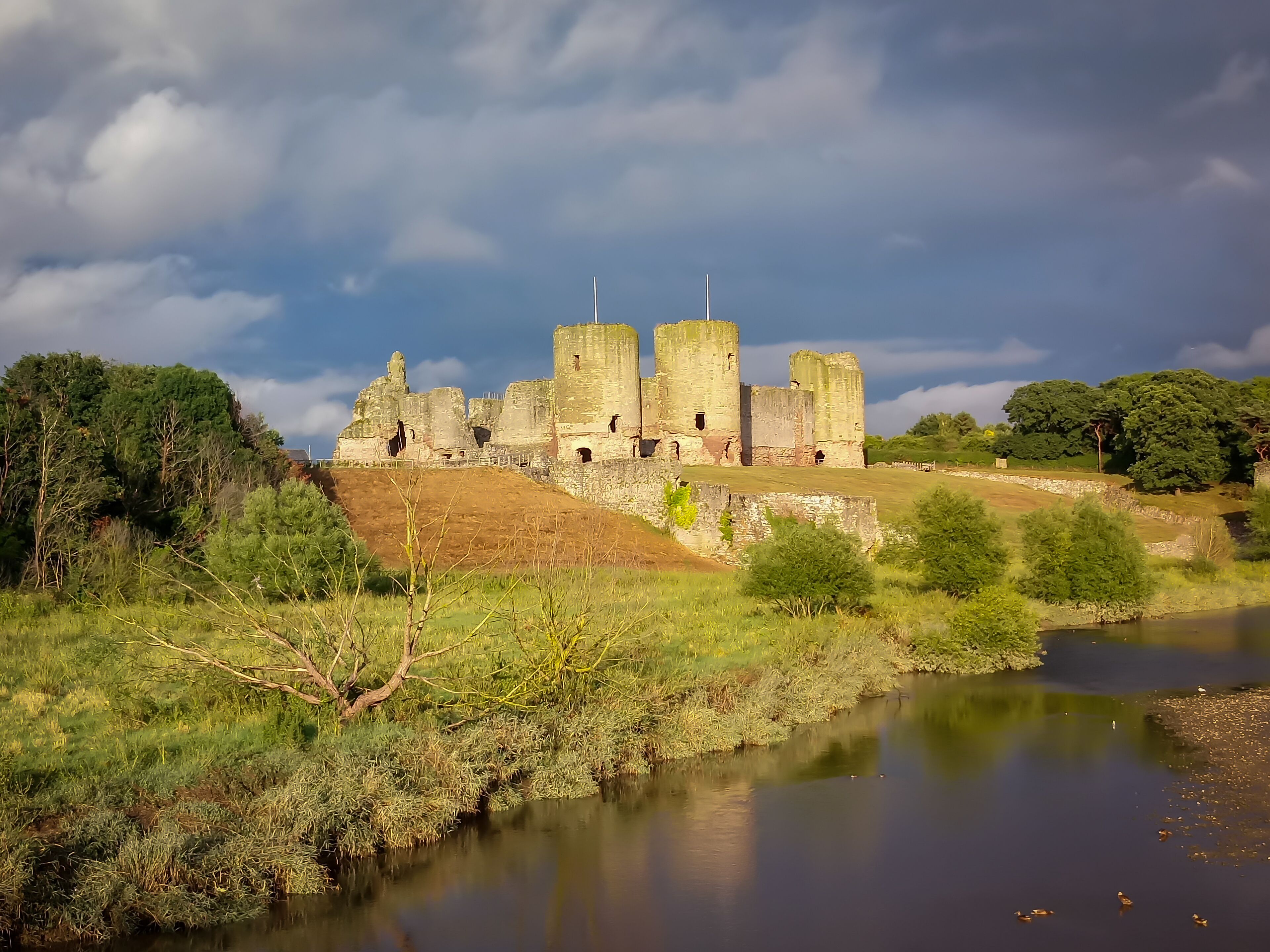 A historic stone castle with towers, surrounded by lush greenery and a calm river.