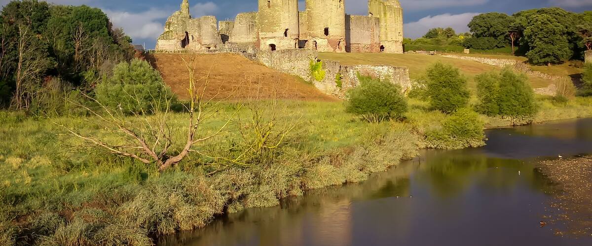 A historic stone castle with towers, surrounded by lush greenery and a calm river.