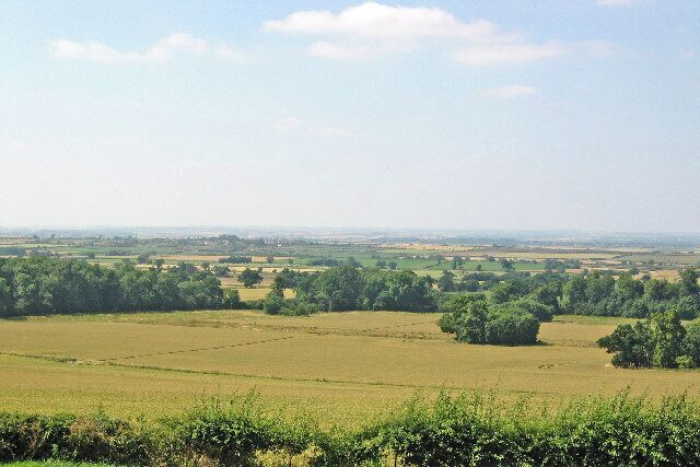 Vale of Belvoir, looking west from Belvoir Castle. Woodland and fields across the near centre of photo are in the square SK8033. The Vale of Belvoir (pronounced Beever) is a large area of agricultural land bounded by long hills, one of which I was standing on when I took this photo. The shallow ridge in the middle distance is typical. The Vale villages are built on these ridges.