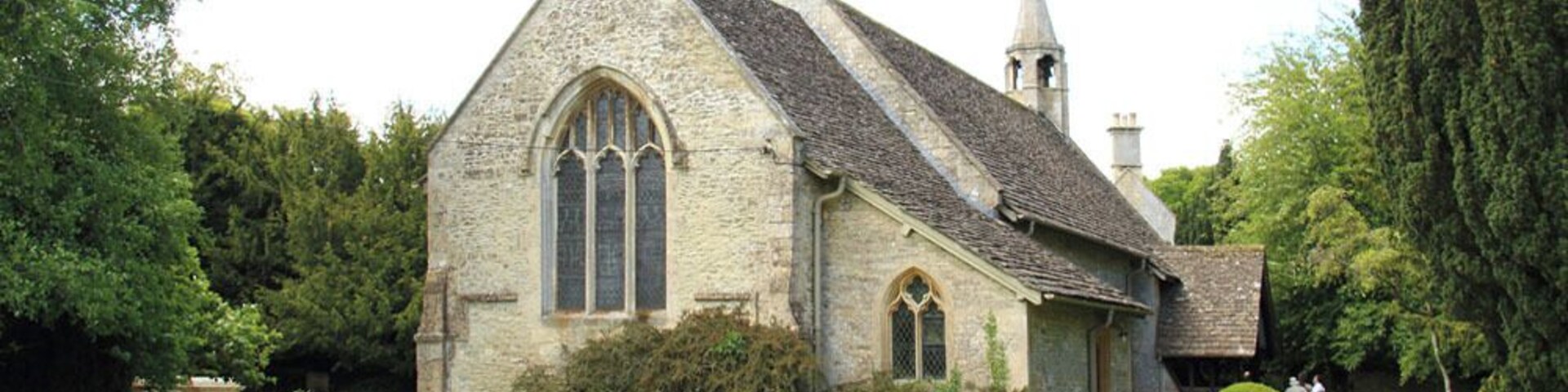 A stone church with a pointed roof and a steeple, surrounded by well-maintained grass and trees.