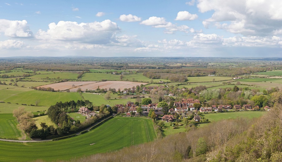A rural landscape with rolling green fields, a small village, and distant hills under a partly cloudy sky.