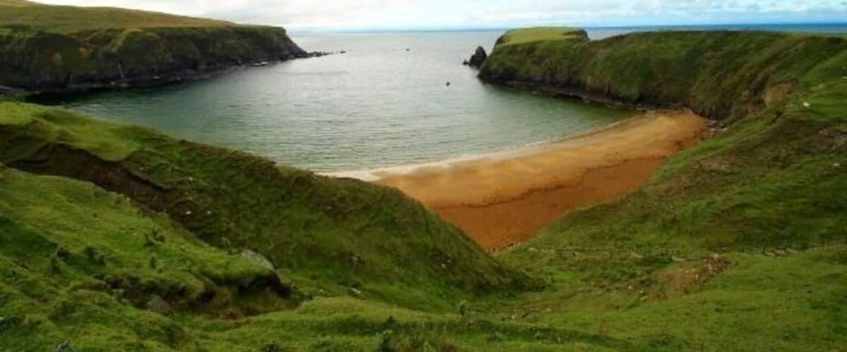 A coastal landscape with a sandy beach, grassy hills, and a calm sea under a cloudy sky.