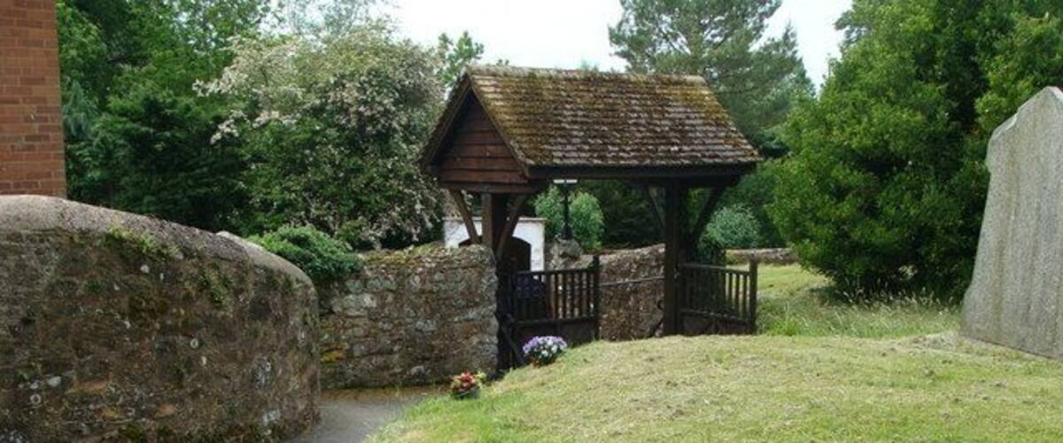 A rural cemetery with a wooden gazebo, a stone wall, and a pathway leading to it.