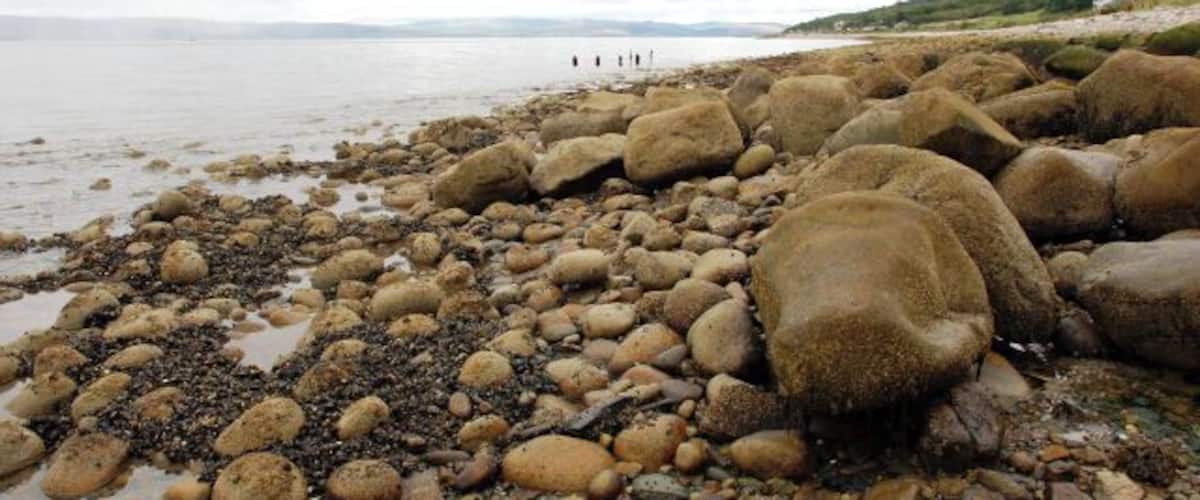 A rocky shoreline with large boulders and pebbles, a calm sea, and a distant view of people and hills.