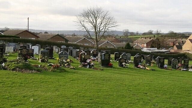 A cemetery with numerous headstones and tombs, surrounded by green grass and residential houses in the background.