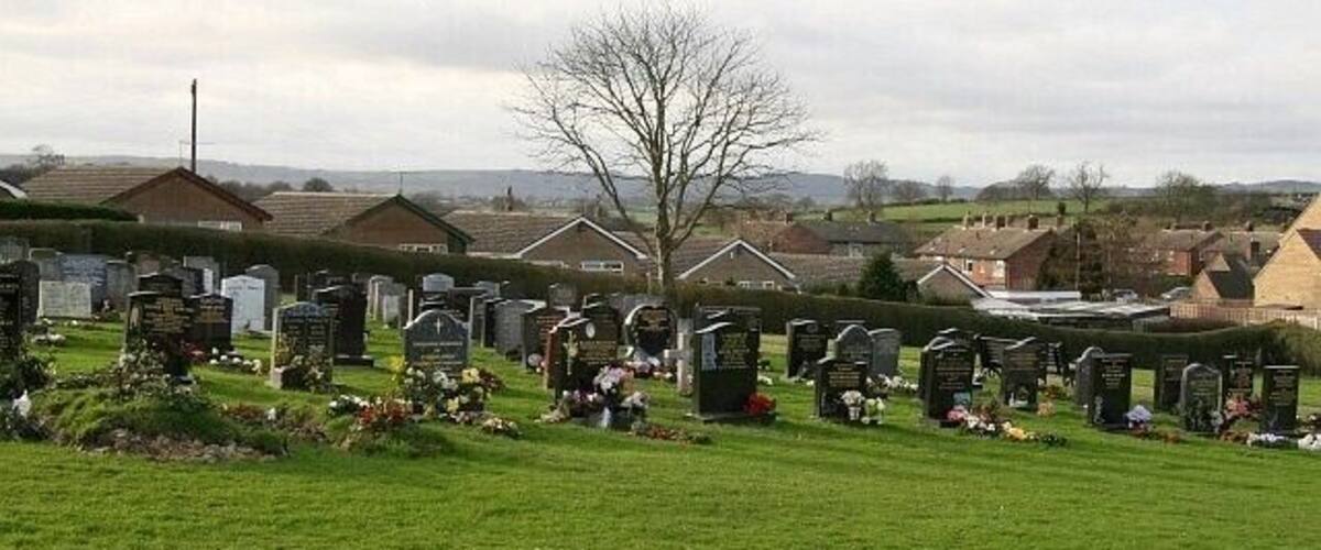 A cemetery with numerous headstones and tombs, surrounded by green grass and residential houses in the background.