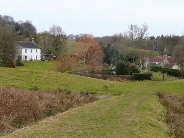 A grassy field with a white house, trees, and a small body of water.