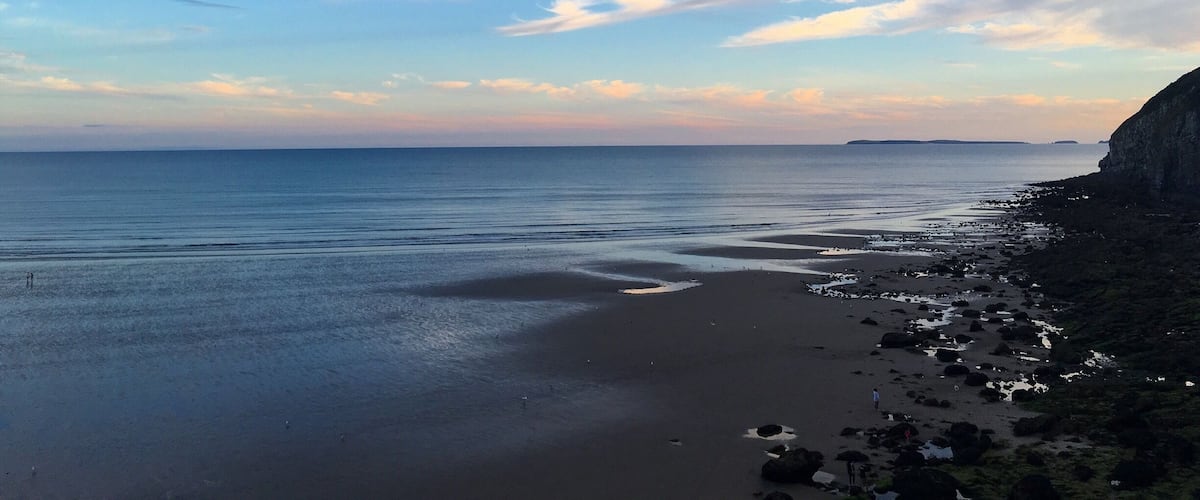 A coastal landscape with a sandy beach, calm sea, and a rocky cliff.