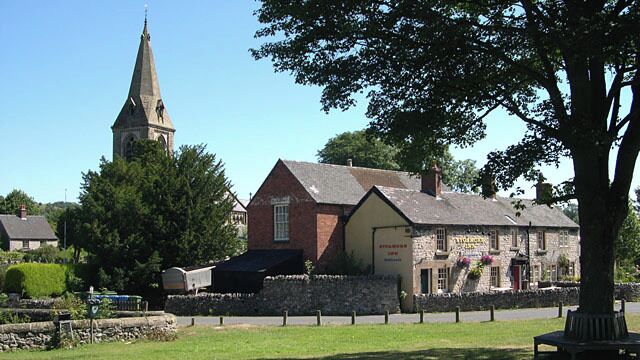 A church with a tall steeple, a stone building with a sign, and a tree in the foreground.