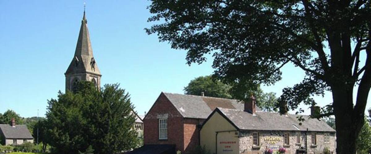 A church with a tall steeple, a stone building with a sign, and a tree in the foreground.
