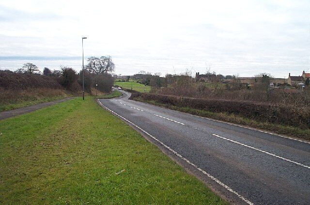 A rural road curving through a green landscape with a lamppost and distant buildings.