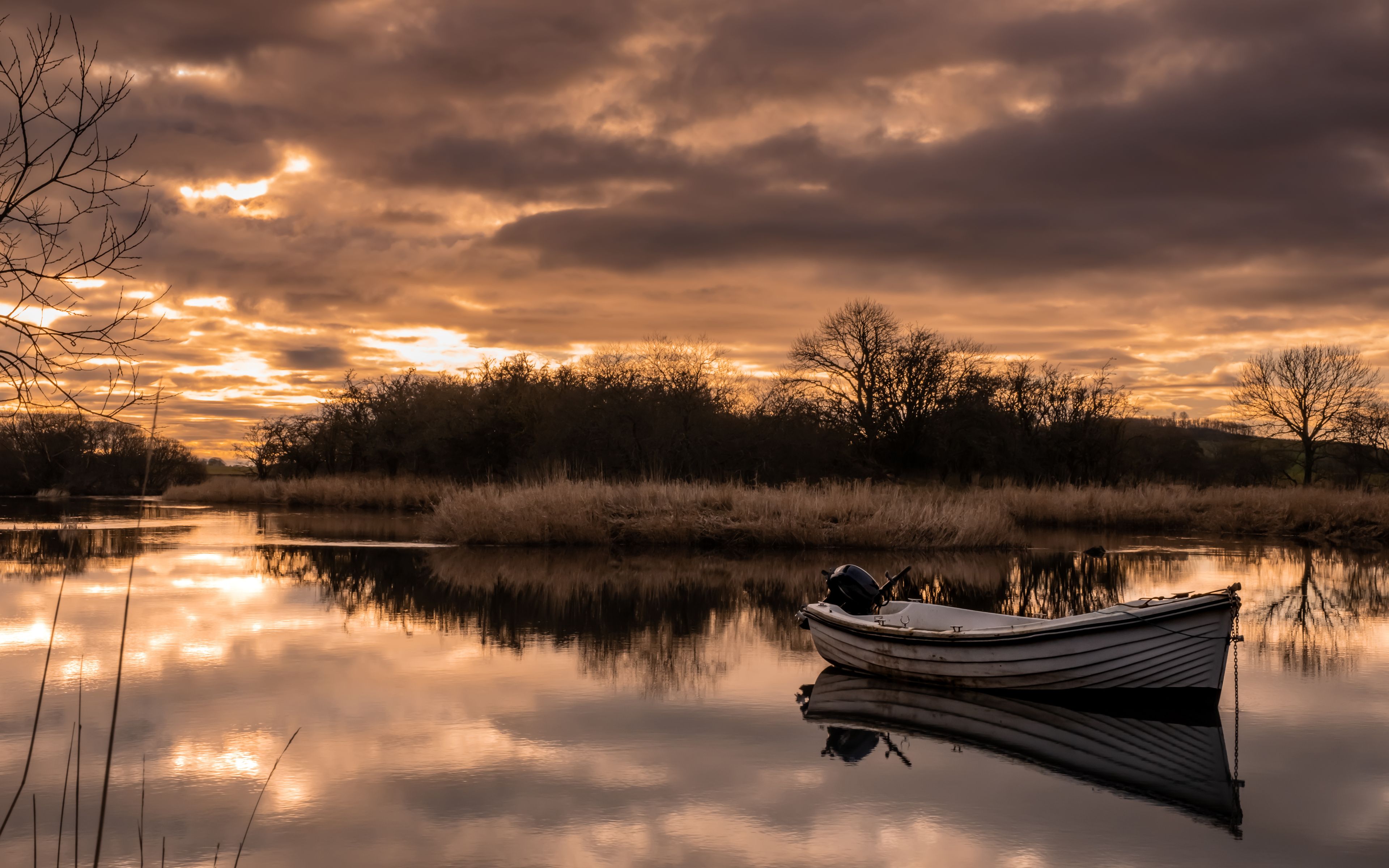 A calm lake with a boat anchored near the shore, surrounded by trees and reeds under a dramatic sunset sky.