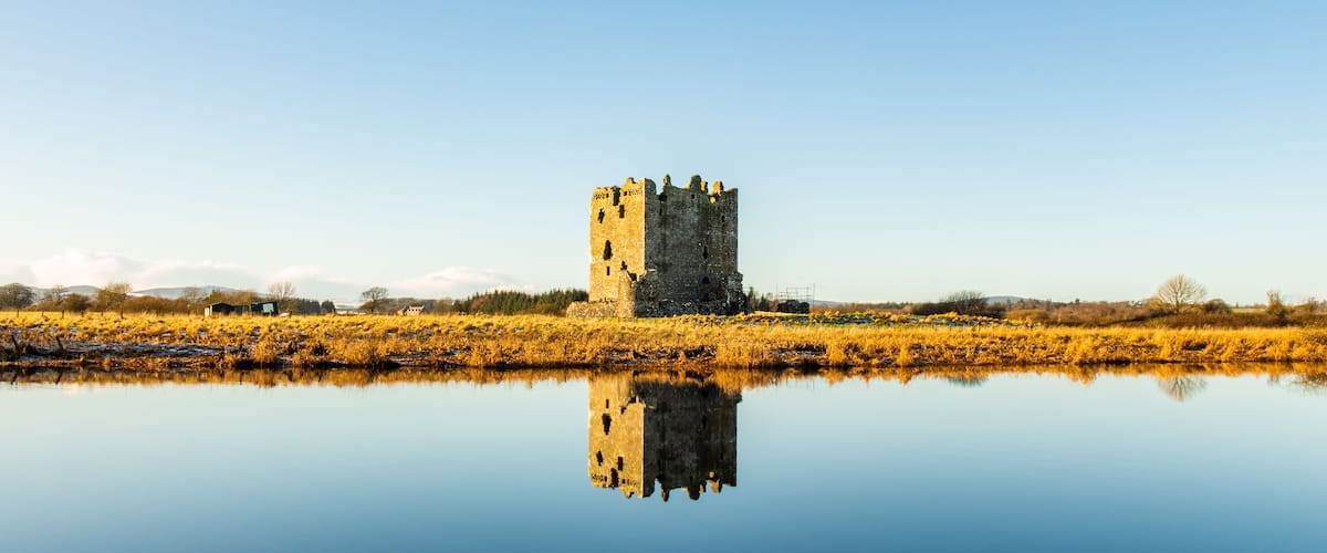 A historic stone tower reflected in a calm body of water, surrounded by a field of yellow flowers and a clear blue sky.