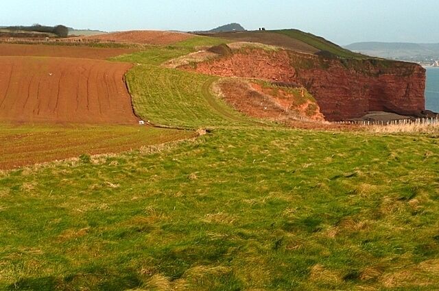 A coastal landscape with a prominent red cliff, green fields, and a distant view of the sea.