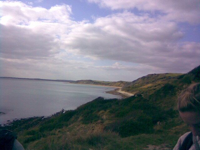Paisagem costeira com praia, colinas gramadas e um corpo d'água sob um céu nublado.