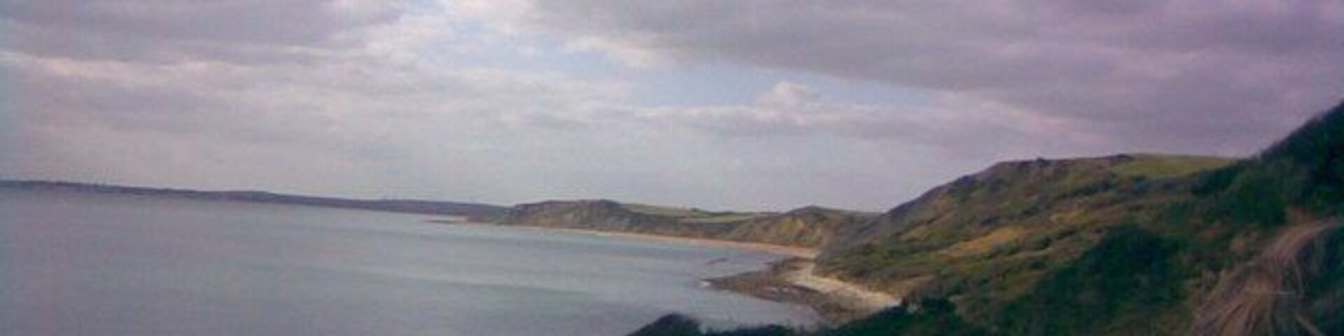 Paisagem costeira com praia, colinas gramadas e um corpo d'água sob um céu nublado.