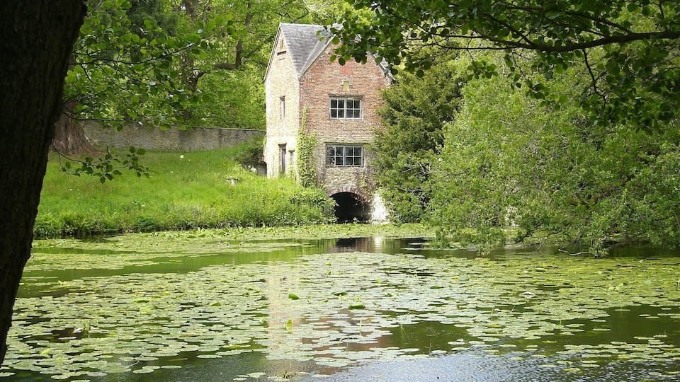 A historic stone building with a brick facade and a small window, surrounded by lush greenery and a pond with lily pads.