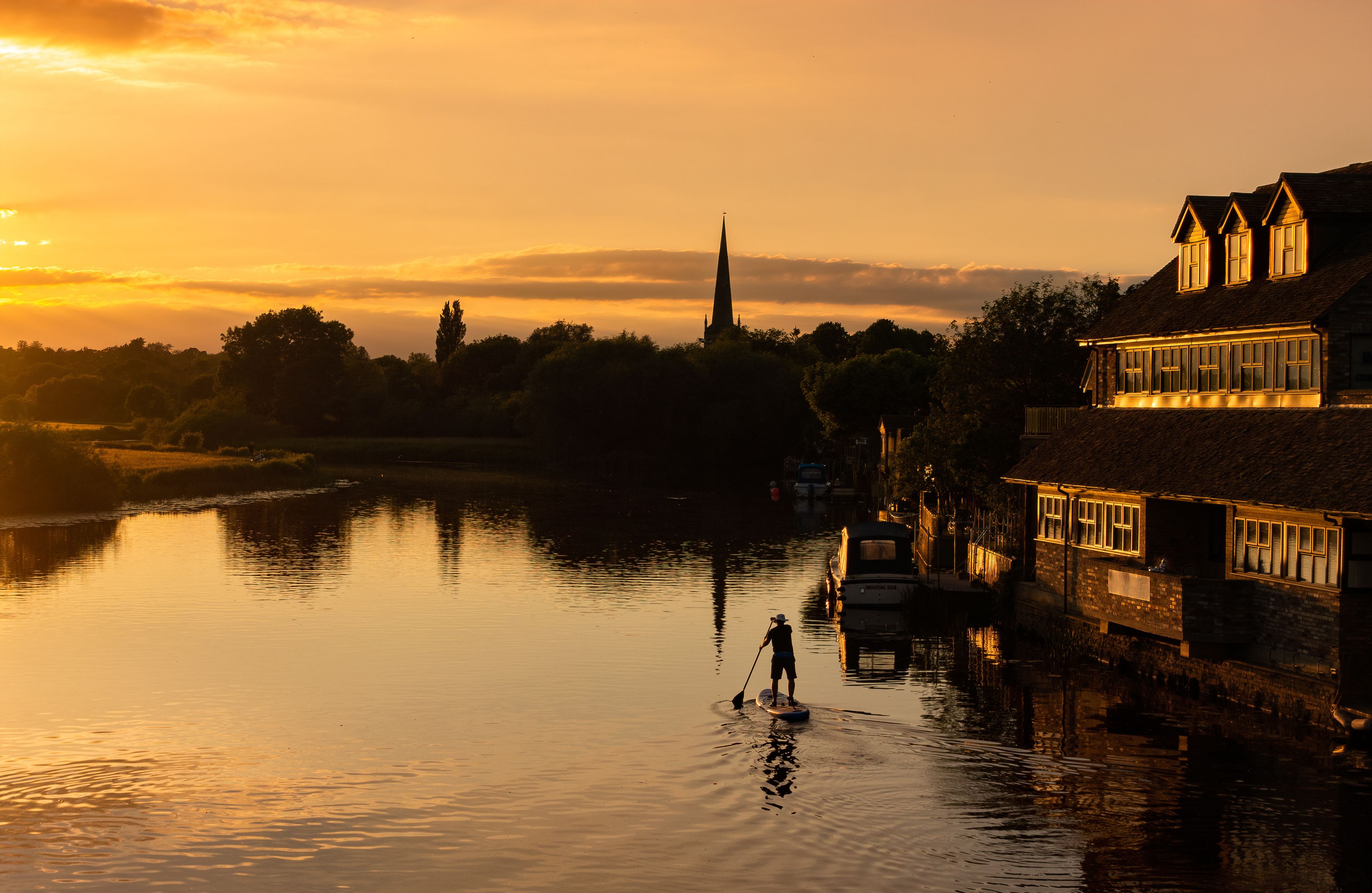 Un atardecer sobre un río con la aguja de una iglesia, barcos y edificios a lo largo de la orilla.