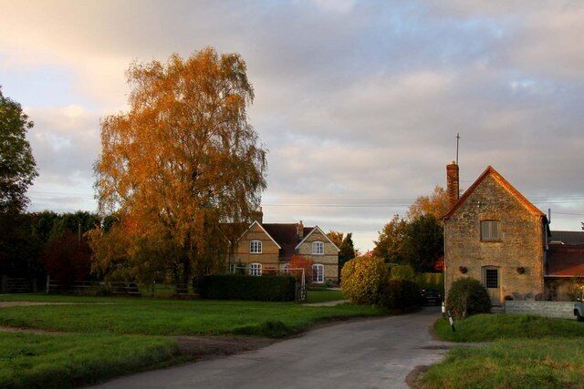A rural road leading to a stone house with a chimney, surrounded by autumn trees and a grassy field.
