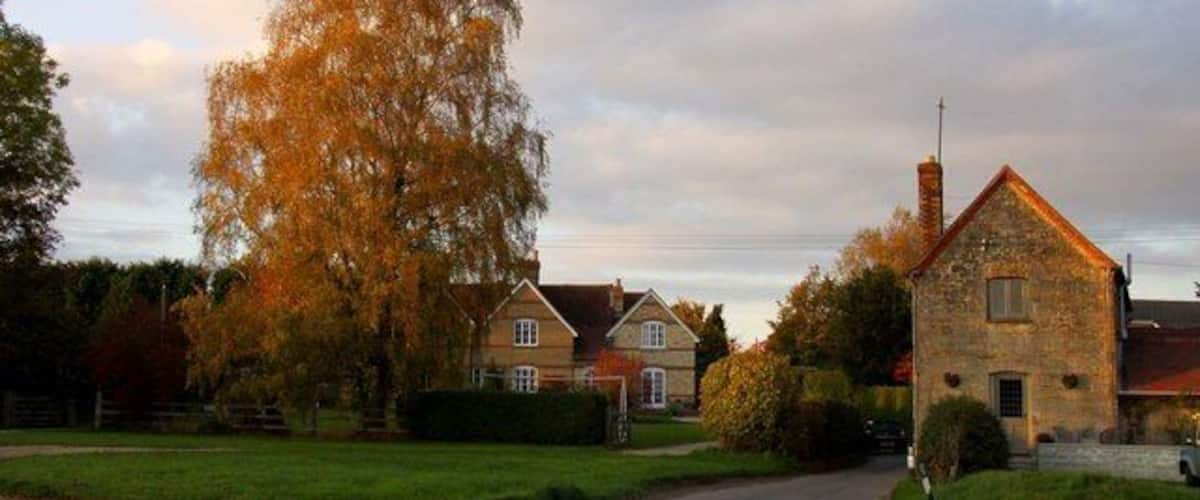 Eine Landstraße führt zu einem Haus aus Stein mit Schornstein, umgeben von Herbstbäumen und einer Wiese.