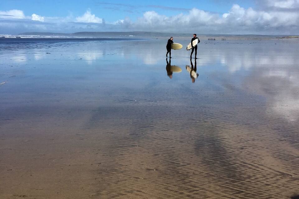Dos surfistes caminen sobre una platja mullada amb ones tranquil·les i un cel serè.
