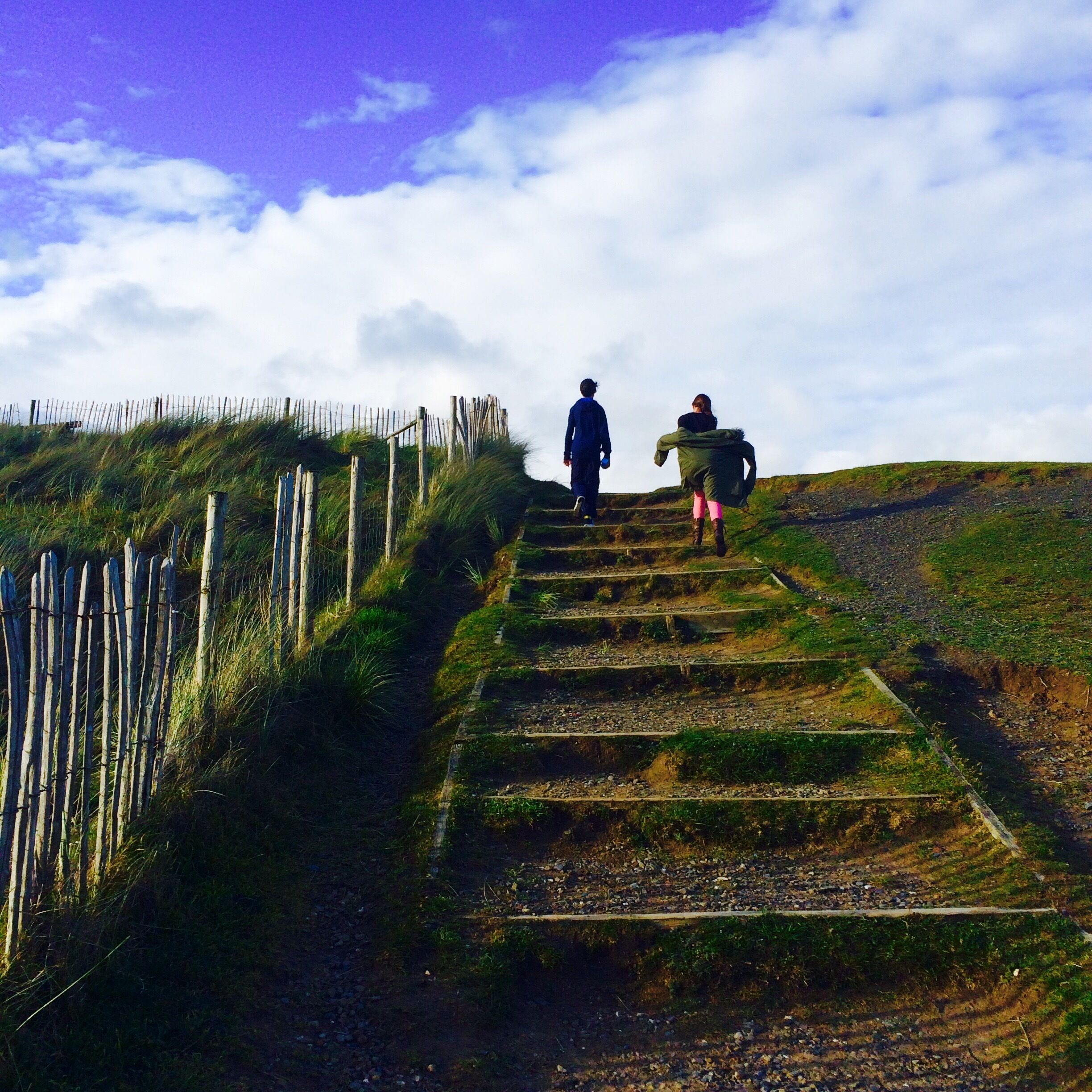 A person walking up a grassy hill with a wooden fence on the left.
