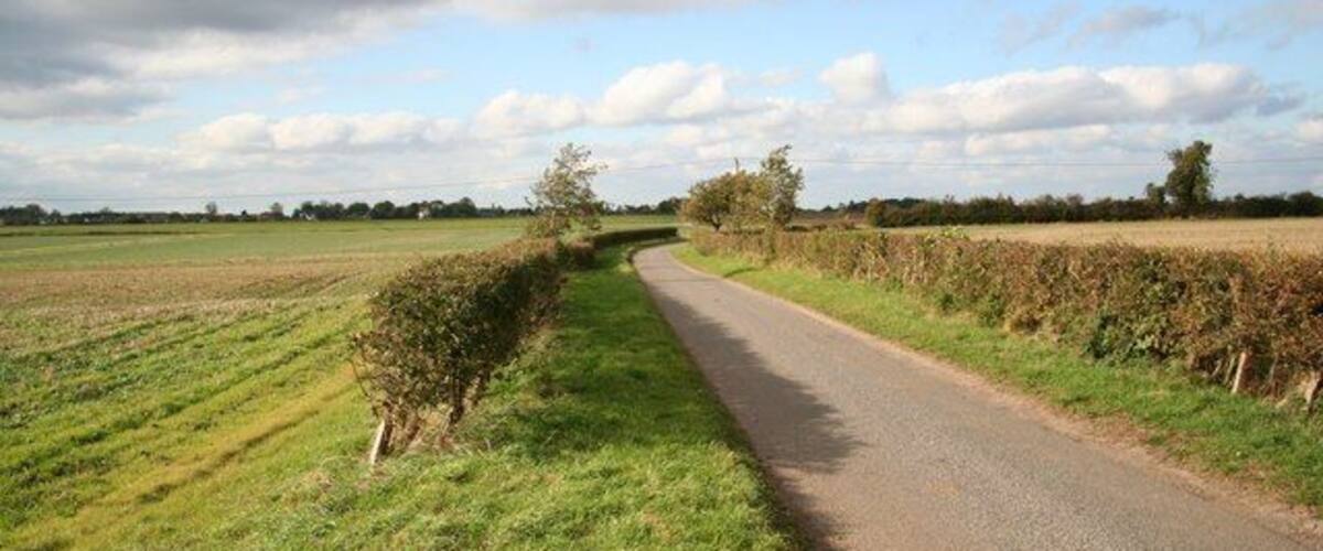A rural road flanked by fields and hedgerows under a partly cloudy sky.