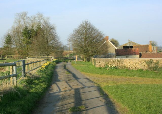 A rural pathway flanked by stone walls and a wooden fence, leading to a house with a red roof.