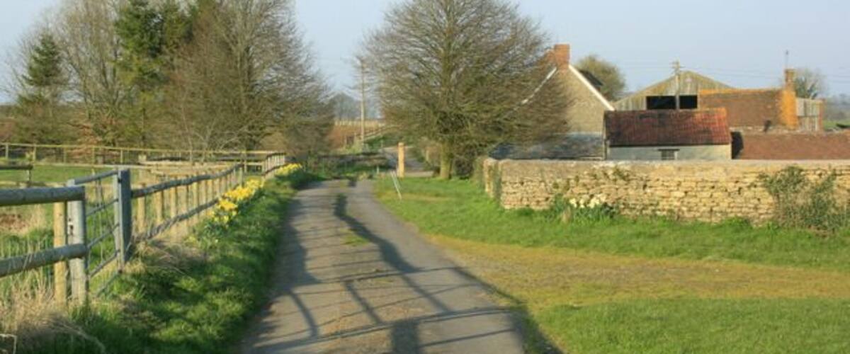 A rural pathway flanked by stone walls and a wooden fence, leading to a house with a red roof.