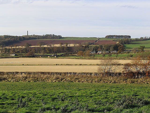 Farmland by Crailing Church A lush pasture field is in the foreground with a stubble field beyond the hedge. Viewed from the northern corner of Crailing Churchyard with the village of Nisbet in the background on the right and the Waterloo Monument at Peniel Heugh on the skyline to the left.