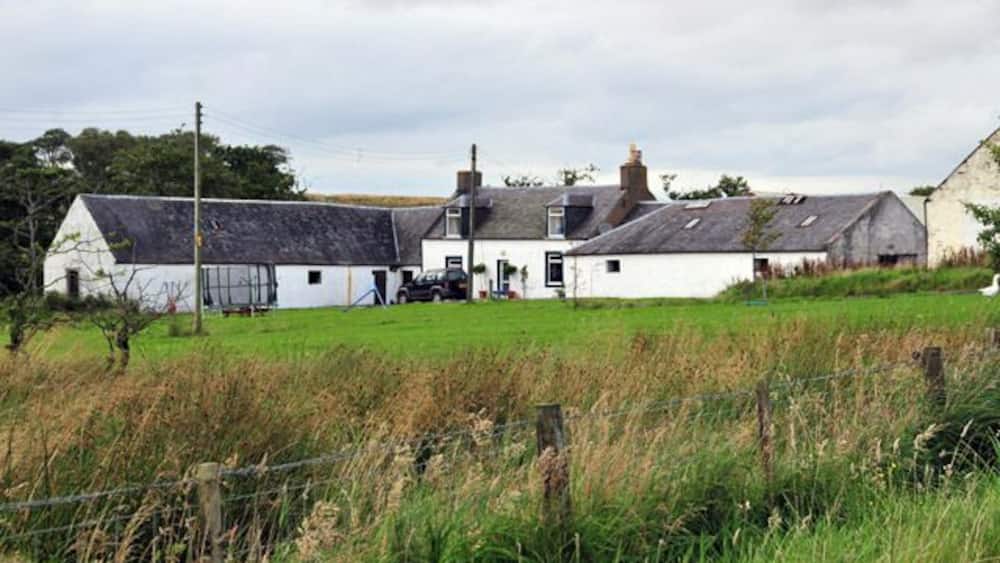 A white farmhouse with a black roof, surrounded by a green field and a fence.