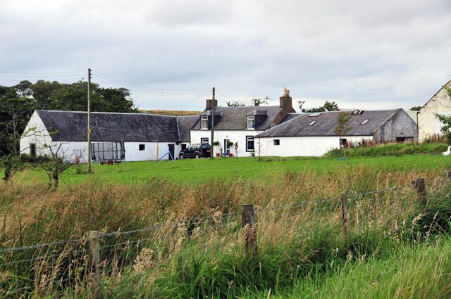 A white farmhouse with a black roof, surrounded by a green field and a fence.