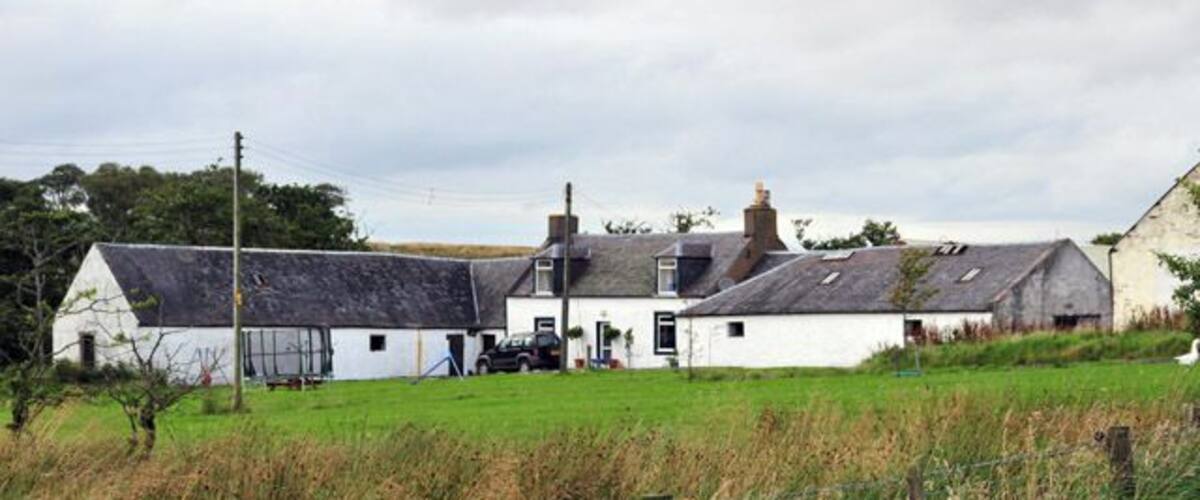 A white farmhouse with a black roof, surrounded by a green field and a fence.