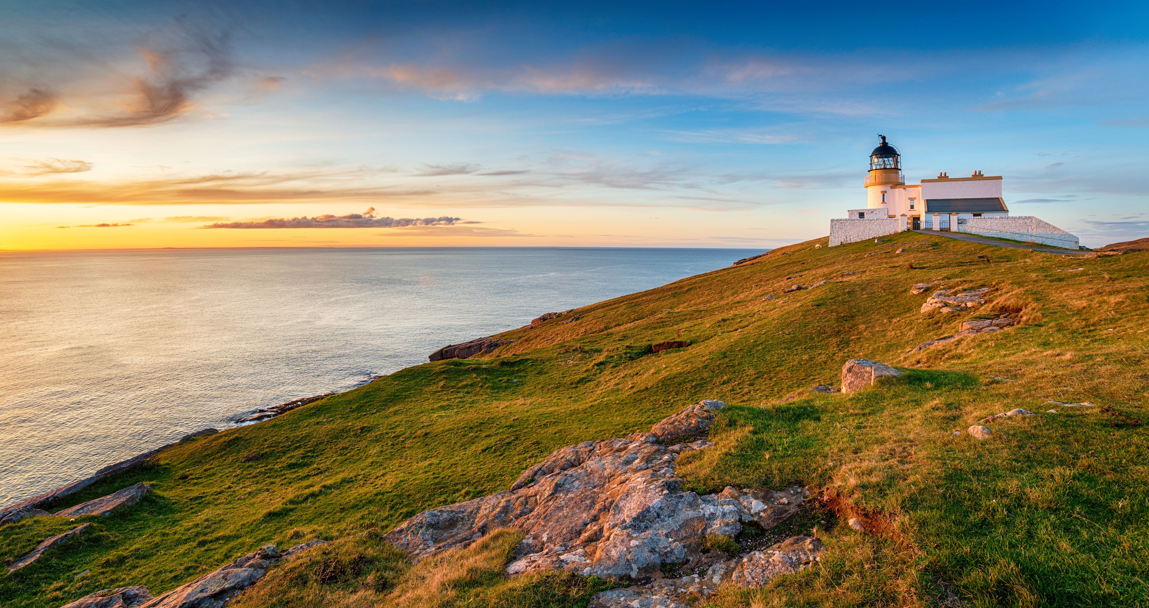 A lighthouse on a grassy hill overlooking the sea at sunset.