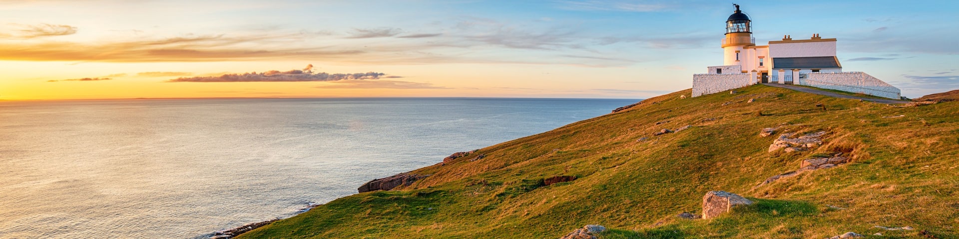 A lighthouse on a grassy hill overlooking the sea at sunset.