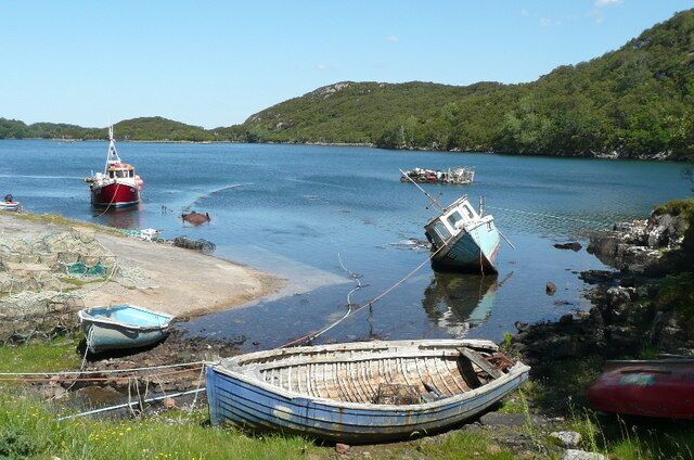 A calm lake with several boats, a rocky shoreline, and green hills in the background.