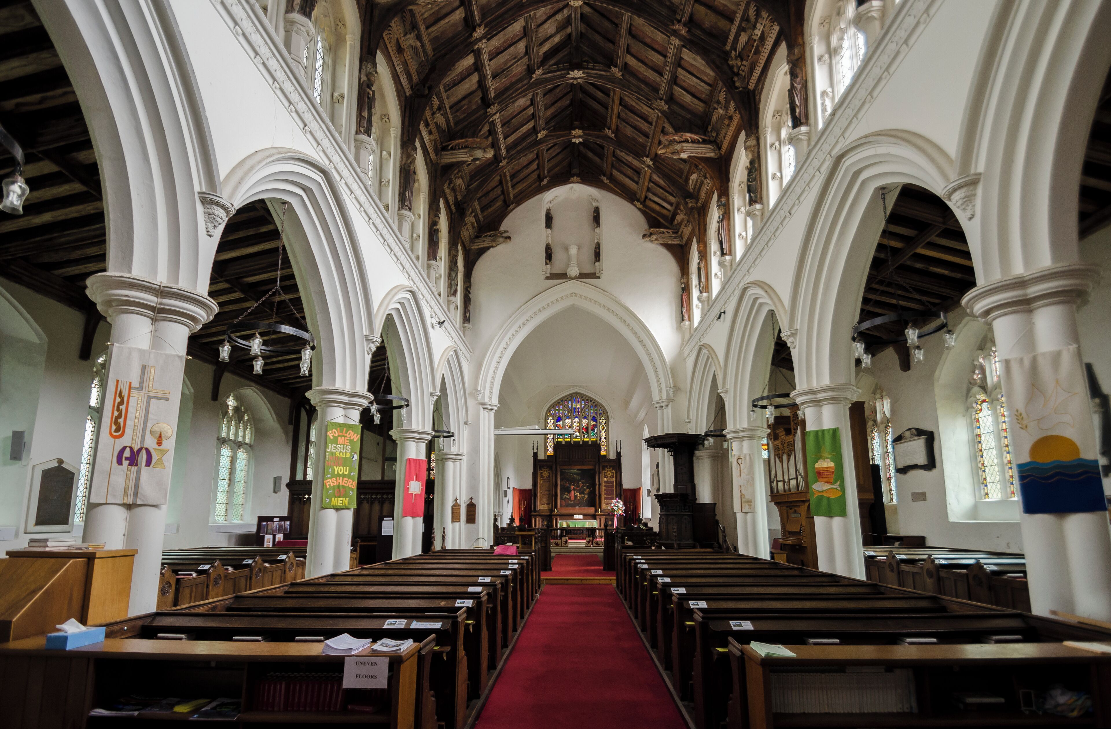 A church interior with wooden pews, a red carpet, white columns, and a wooden ceiling.