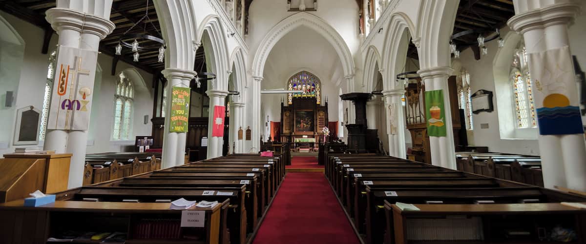 A church interior with wooden pews, a red carpet, white columns, and a wooden ceiling.