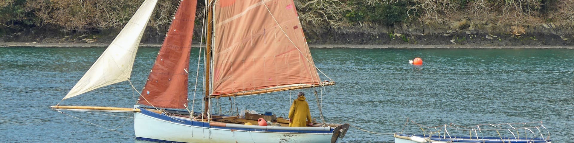 A sailboat with two sails on a calm lake, a smaller boat nearby, and a forested shoreline in the background.