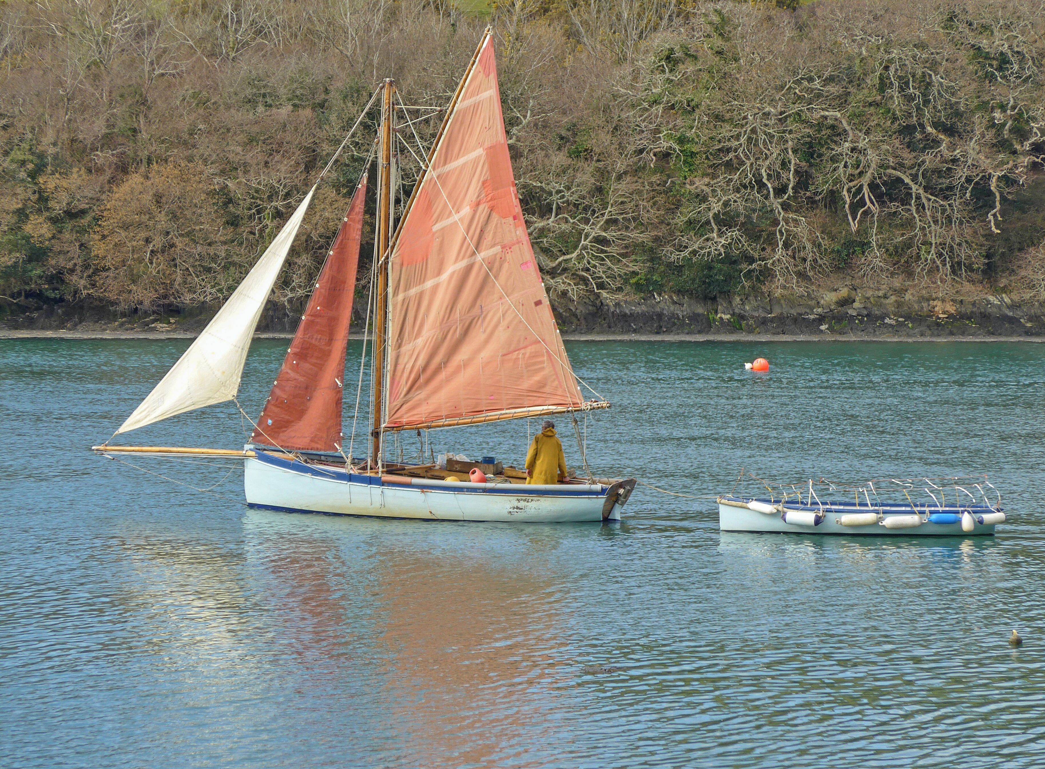 A sailboat with two sails on a calm lake, a smaller boat nearby, and a forested shoreline in the background.