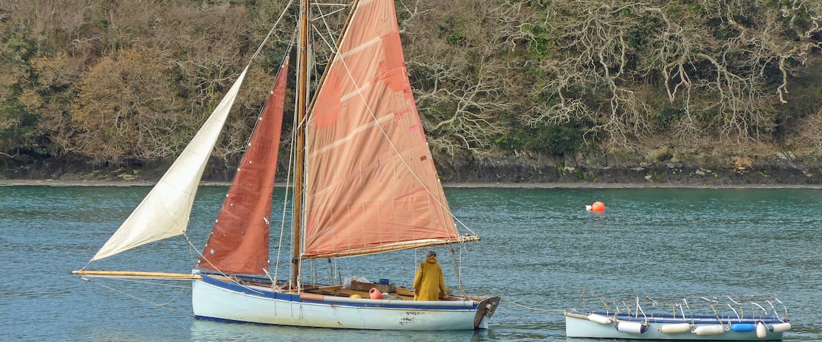 A sailboat with two sails on a calm lake, a smaller boat nearby, and a forested shoreline in the background.