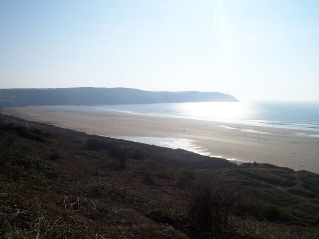 A coastal landscape with a beach, grassy hills, and a distant shoreline.