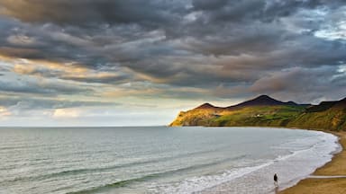 Une plage où une personne se promène le long du rivage, une montagne en arrière-plan et un ciel nuageux.