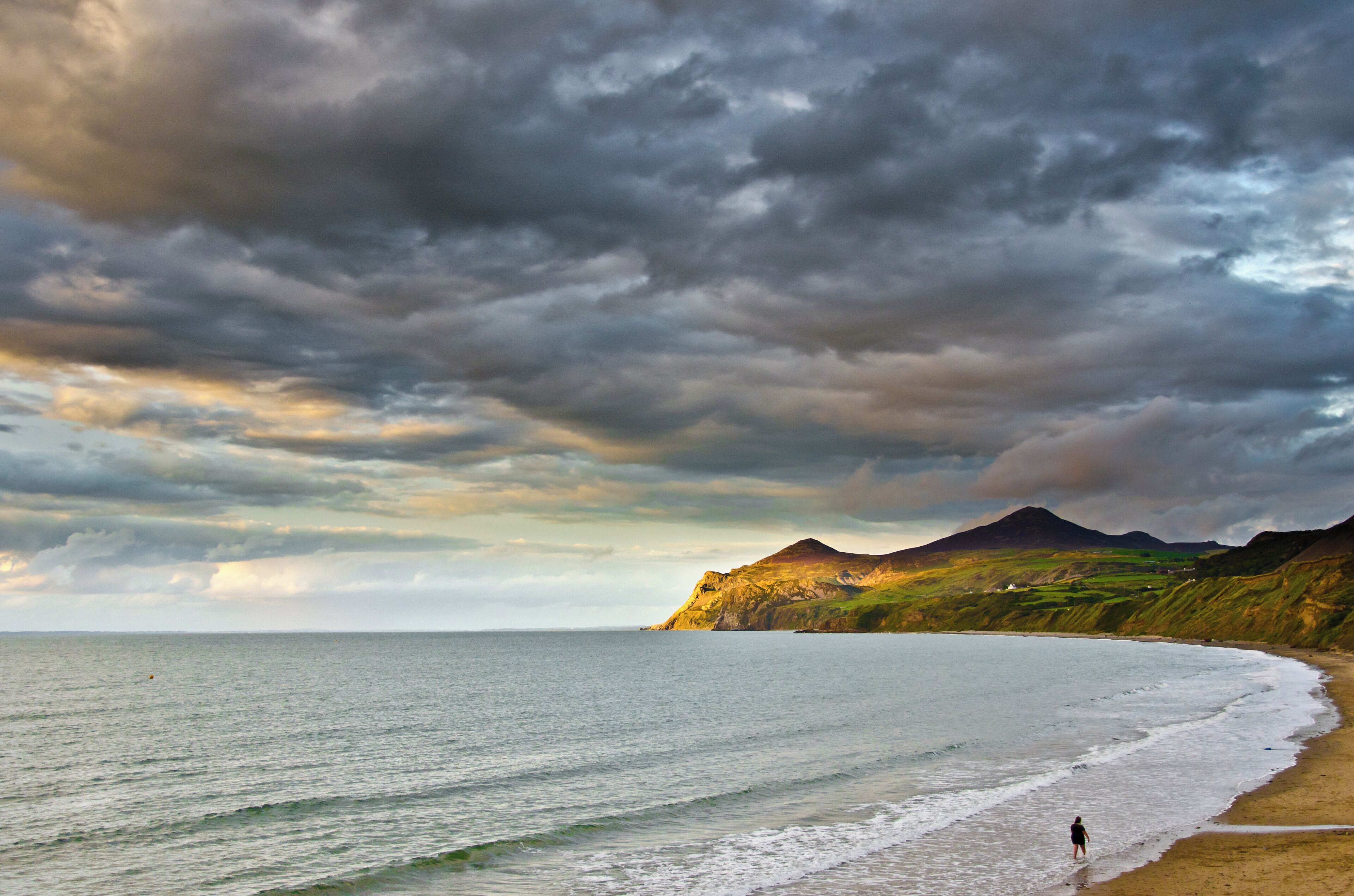 A beach with a person walking along the shore, a mountain in the background, and a cloudy sky.