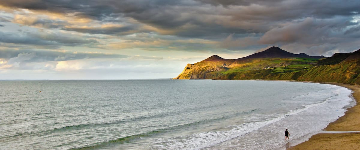 A beach with a person walking along the shore, a mountain in the background, and a cloudy sky.